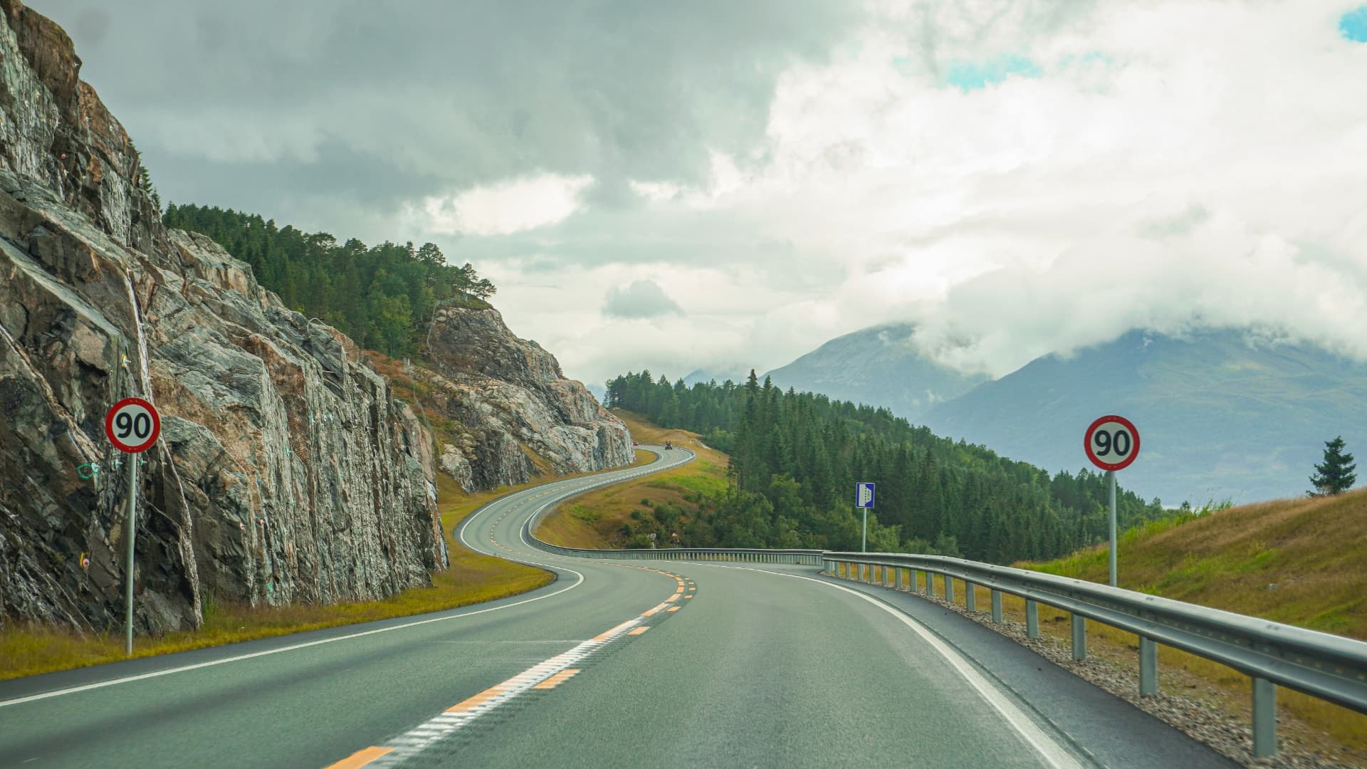 Winding mountain road with rocky cliffs and forested peaks under a dramatic sky